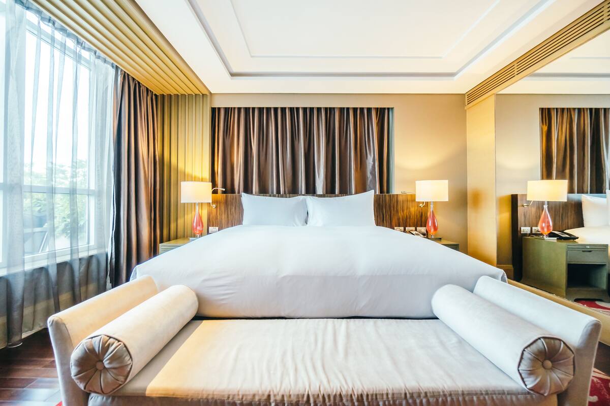A large bed with white linens, two bedside tables with lamps, and a view of the outdoors through sheer curtains.