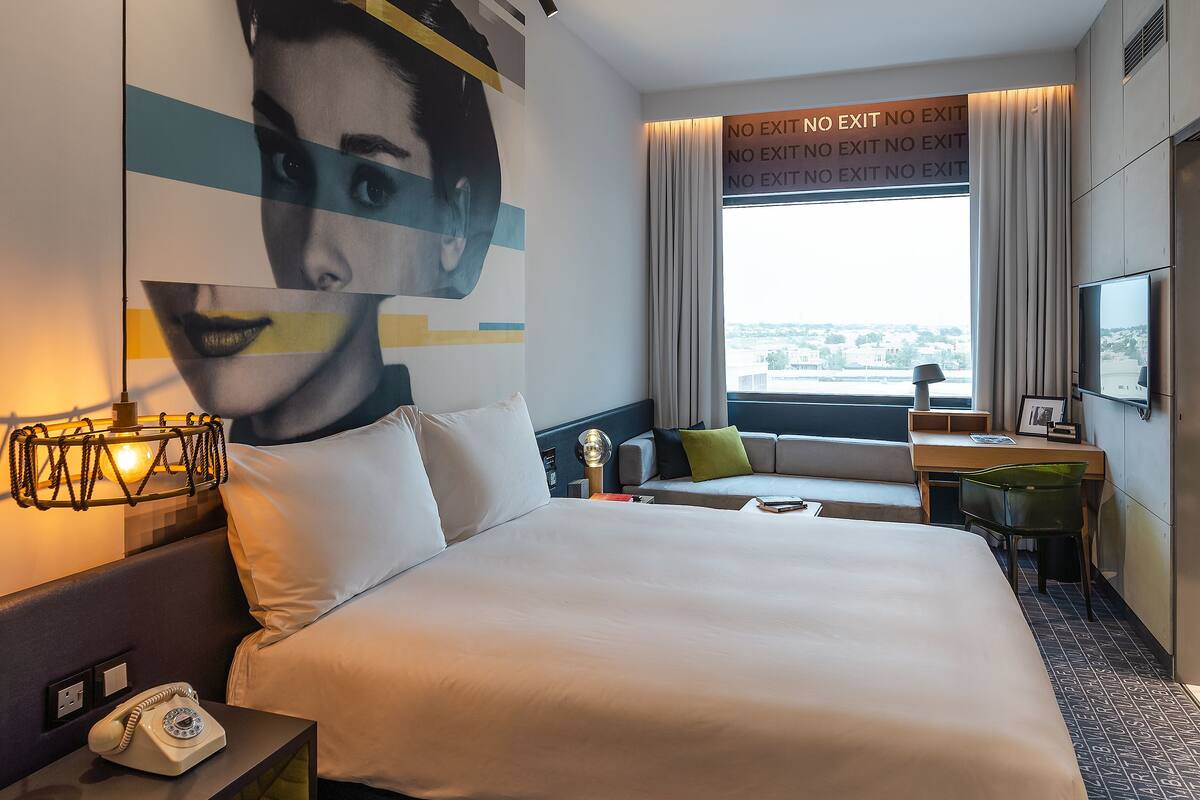 Ein modernes Hotelzimmer mit einem großen Bett, einem Schreibtisch, einem Sessel, einem Fernseher und Blick auf die Stadt.