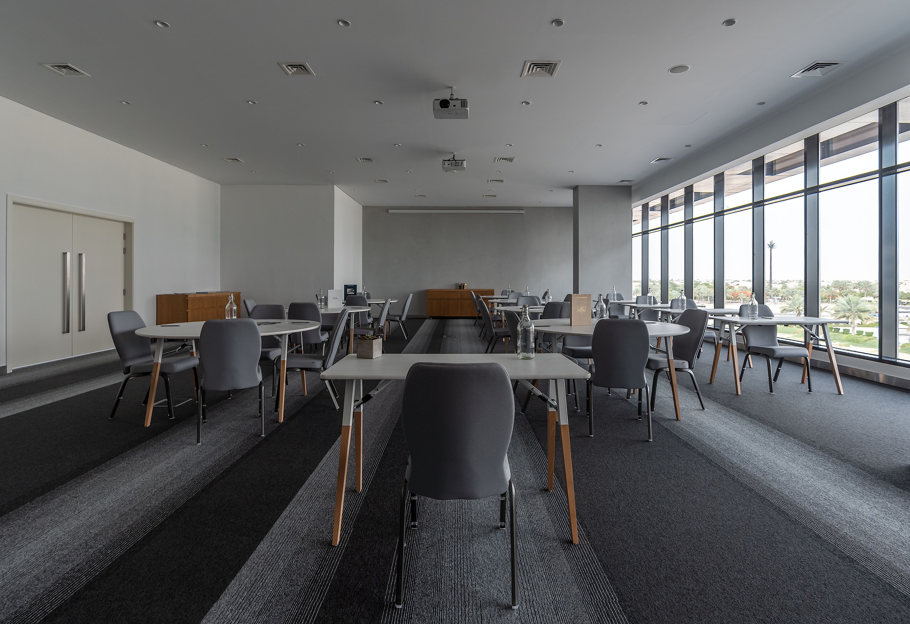 A modern conference room with tables and chairs, large windows, and a projector on the ceiling.