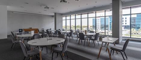 Una sala de conferencias moderna con varias mesas y sillas, ventanas amplias con vista a la ciudad y un proyector en el techo.