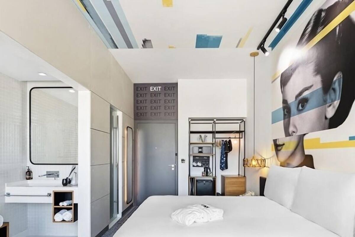 Ein modernes Hotelzimmer mit einem großen Bett, einem Badezimmer mit Spiegel und Waschbecken sowie einem Kleiderschrank. An der Wand befinden sich mehrere Fluchtzeichen.