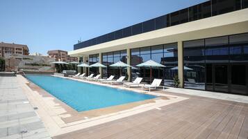 A swimming pool with lounge chairs and umbrellas, surrounded by a wooden deck and modern buildings.