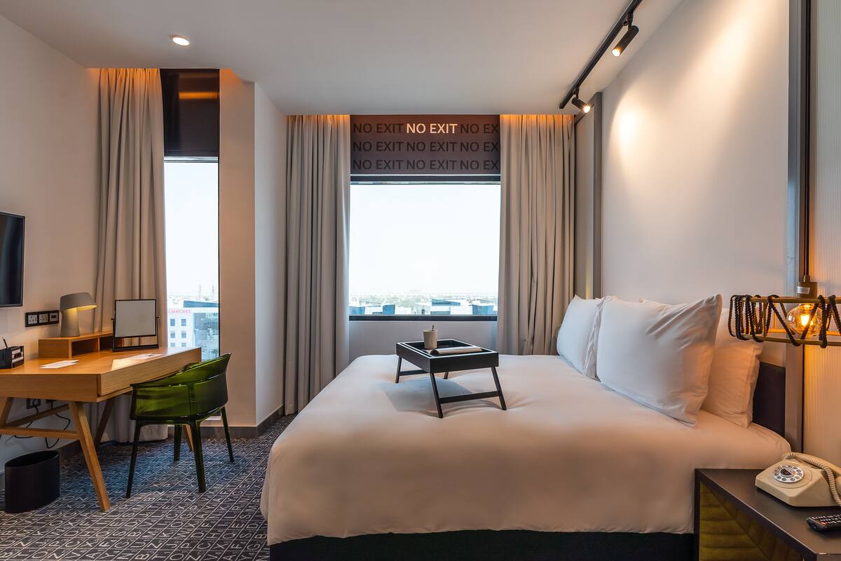Ein Hotelzimmer mit einem Bett, einem Schreibtisch mit Stuhl, einem Nachttisch mit Lampe und einem Fenster mit Vorhängen.