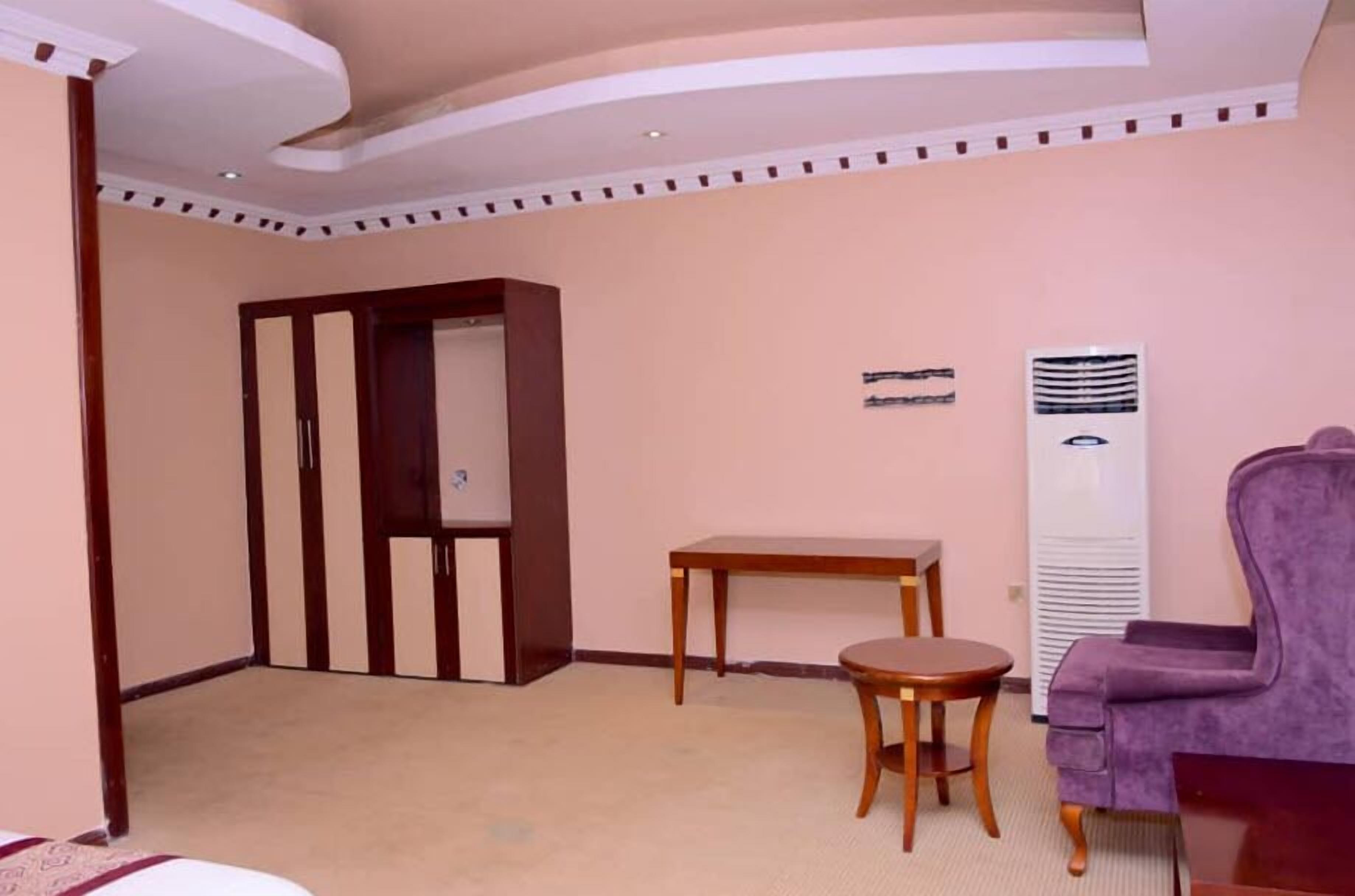 A hotel room with a wooden wardrobe, a small round table, a purple armchair, and a standing heater.