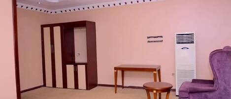 A hotel room with a wooden wardrobe, a small round table, a purple armchair, and a standing heater.