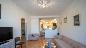 A living room with a TV, seating, a glass coffee table, and a kitchen in the background. - Meidan Suites (Tbilisi)