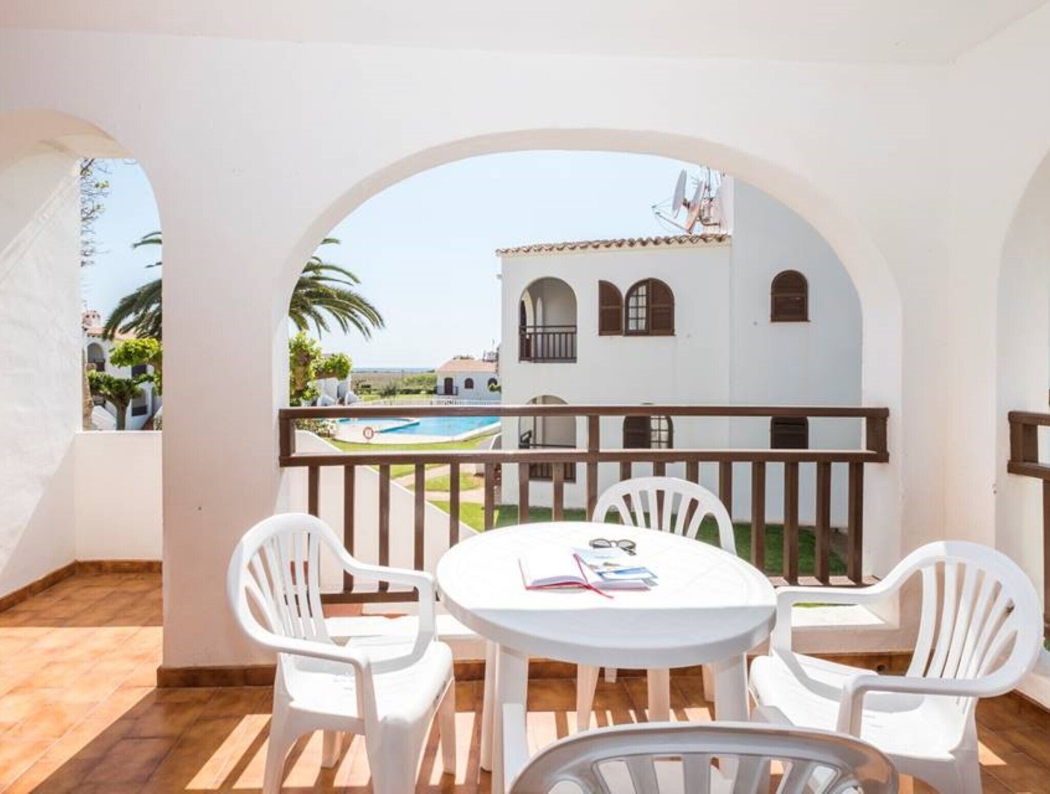 A balcony with white plastic chairs and a table, overlooking a swimming pool and a white building with arched windows.