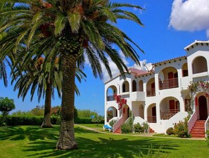 A white building with multiple balconies and arches, surrounded by well-maintained grass and palm trees.