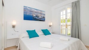 A hotel room with a bed, two pillows, a painting of boats, a ceiling fan, and a window with curtains. - Apartment in Well-Maintained Complex with Pool - Girasol Garden Superior 2 F (menorca)