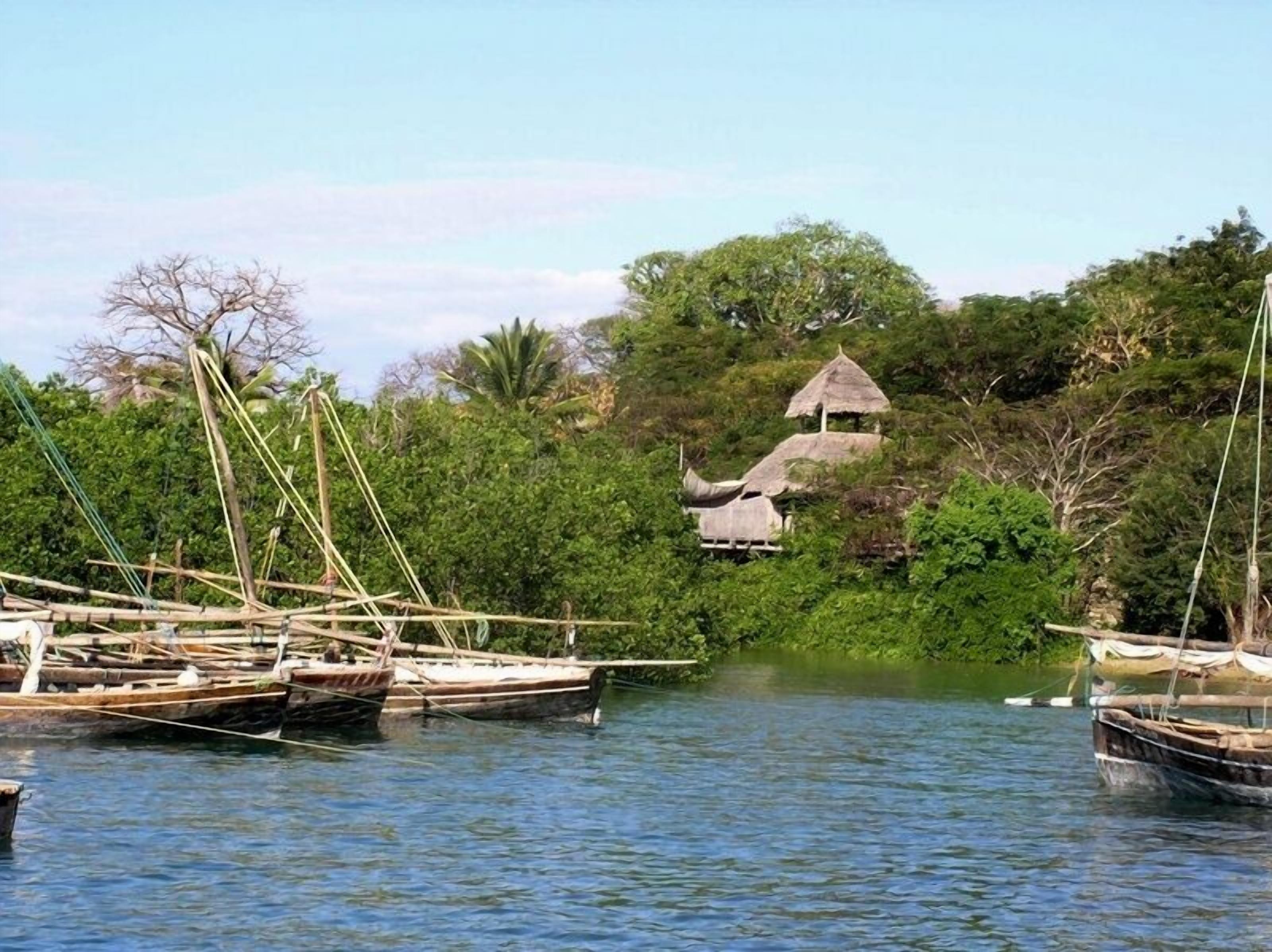 Un río tranquilo con barcas tradicionales amarradas a la izquierda. Al fondo, se observa una pequeña construcción de techo de paja rodeada de abundante vegetación.