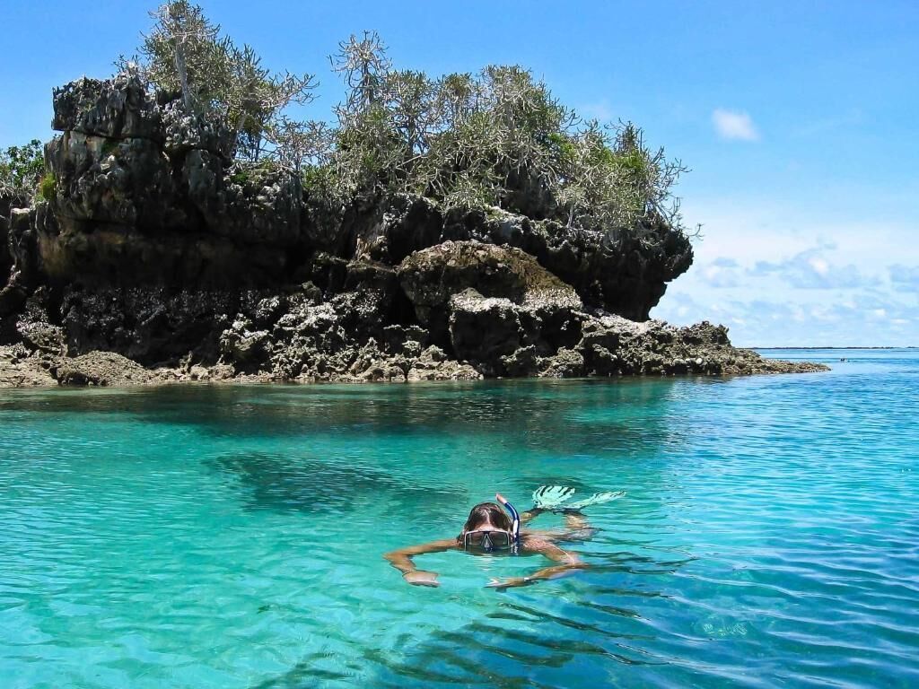 A person snorkeling in clear turquoise water with rocky cliffs and sparse vegetation in the background.