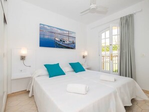 A hotel room with a bed, two pillows, a painting of boats, a ceiling fan, and a window with curtains.