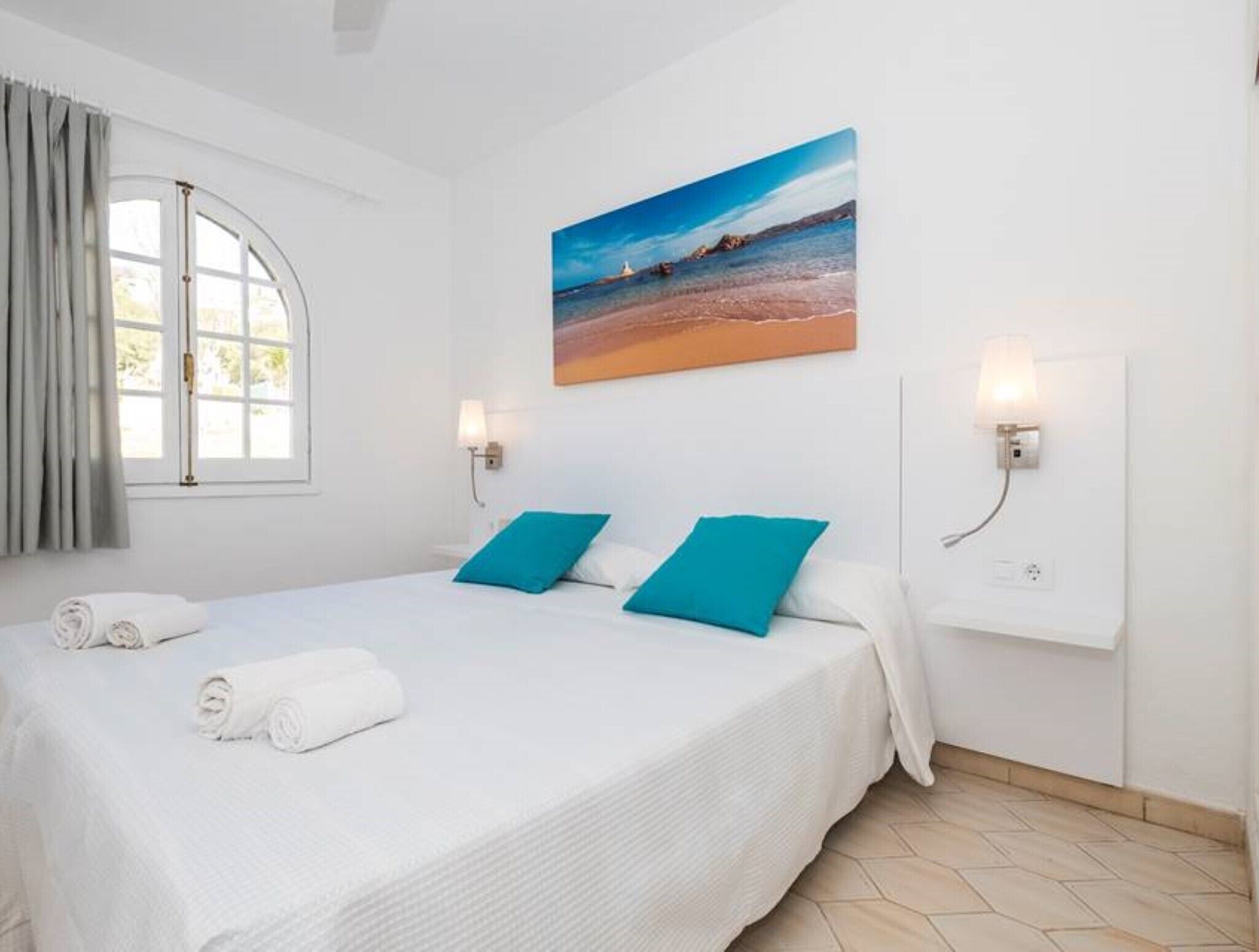 Een hotelkamer met een bed, twee kussens, een strandafbeelding en twee wandlampen.