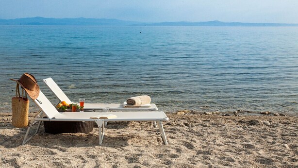 A beach chair with a hat and a basket, a towel, and a glass of drink on the sand near the water.