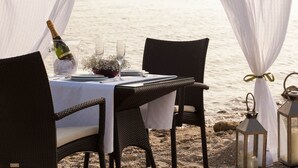 A beachside dining setup with wicker chairs, a table set with wine glasses, a bottle of champagne, and lanterns.