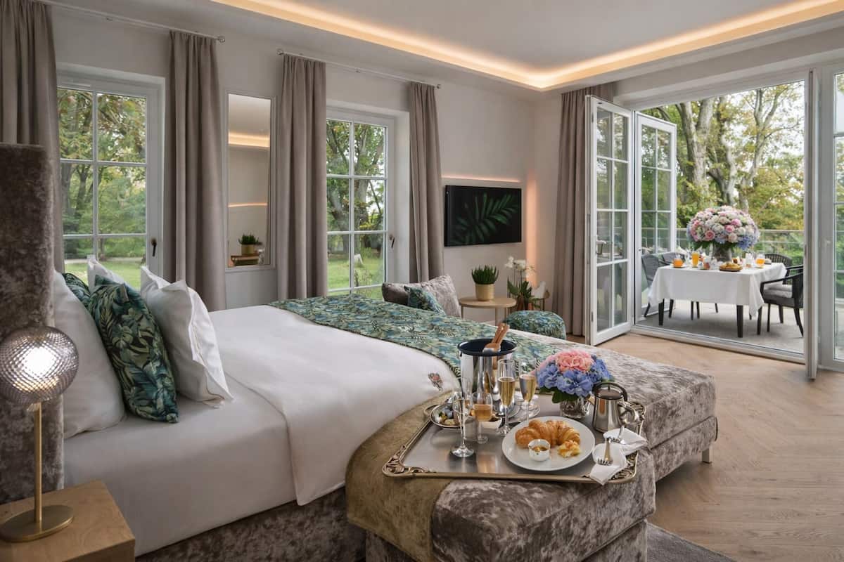 Une chambre spacieuse avec un grand lit, un coin repas avec une table dressée pour deux personnes, et une vue sur l’extérieur.
