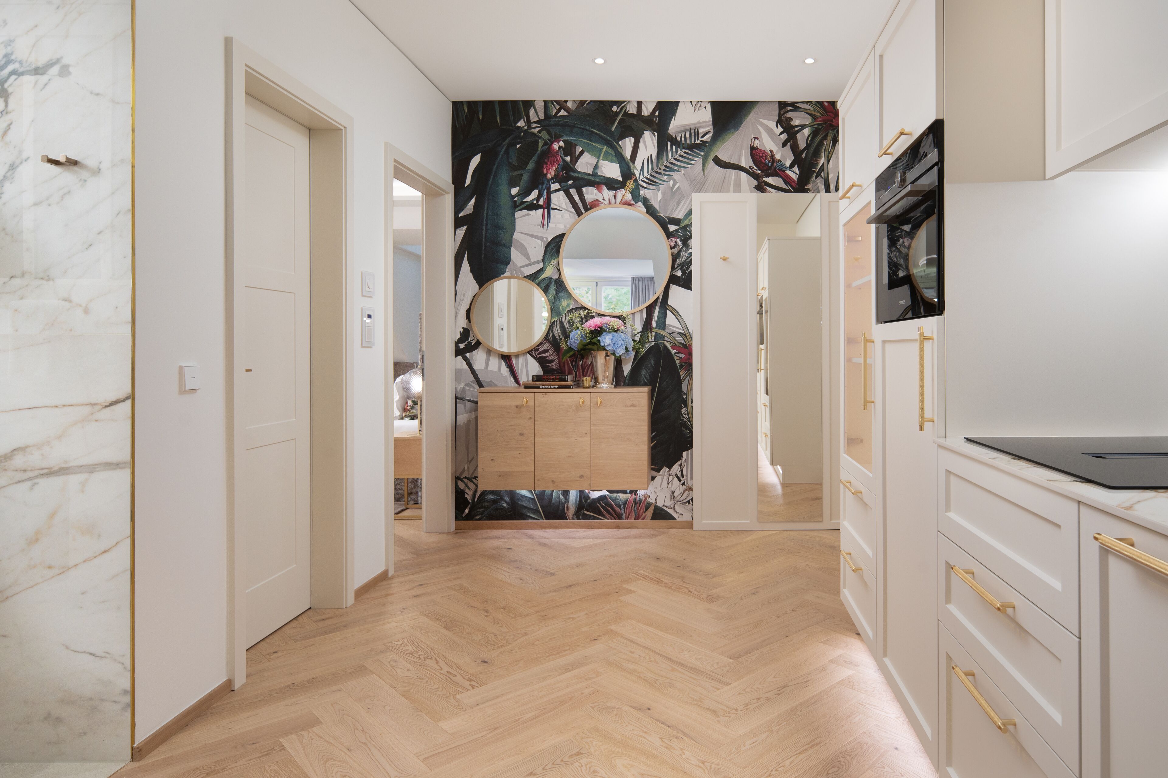 A hallway with a herringbone floor, white walls, and a wooden cabinet with gold handles.