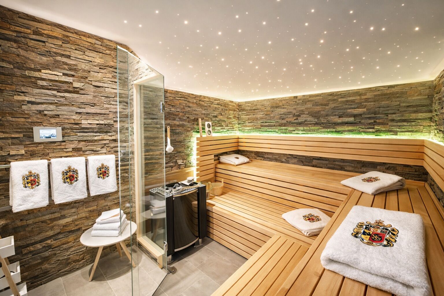 Une salle de spa moderne avec un mur en pierre, des sièges en bois, une douche en verre et des serviettes ornées d’un motif héraldique.
