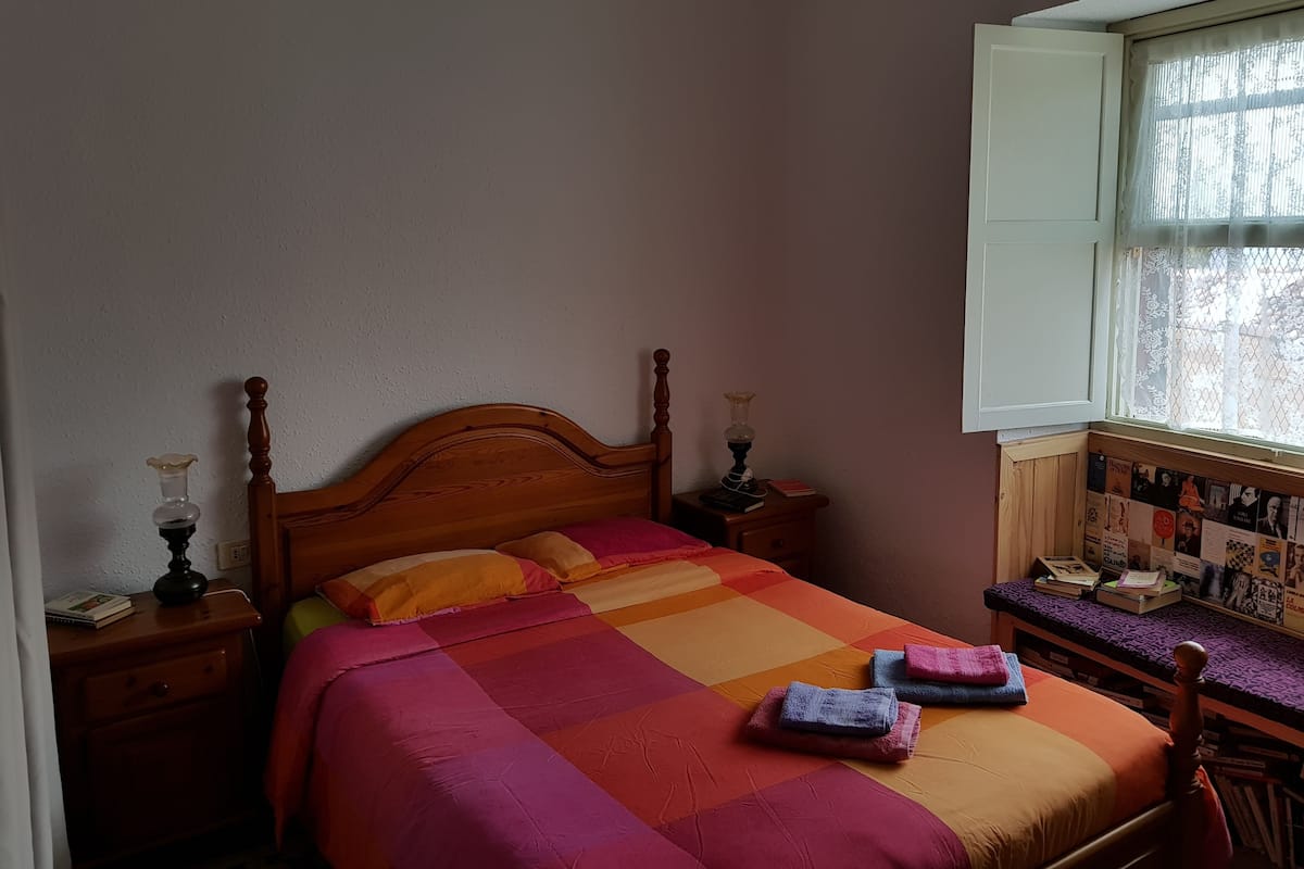A bedroom with a wooden bed, a nightstand with a lamp, a window with lace curtains, and a wall with a bulletin board.