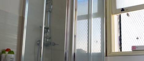 A glass-enclosed shower stall with a white tiled floor and a window with blinds.