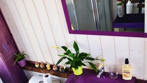 Une salle de bain avec un lavabo violet, un miroir au-dessus, des toilettes et une petite étagère avec des plantes et des articles de toilette.