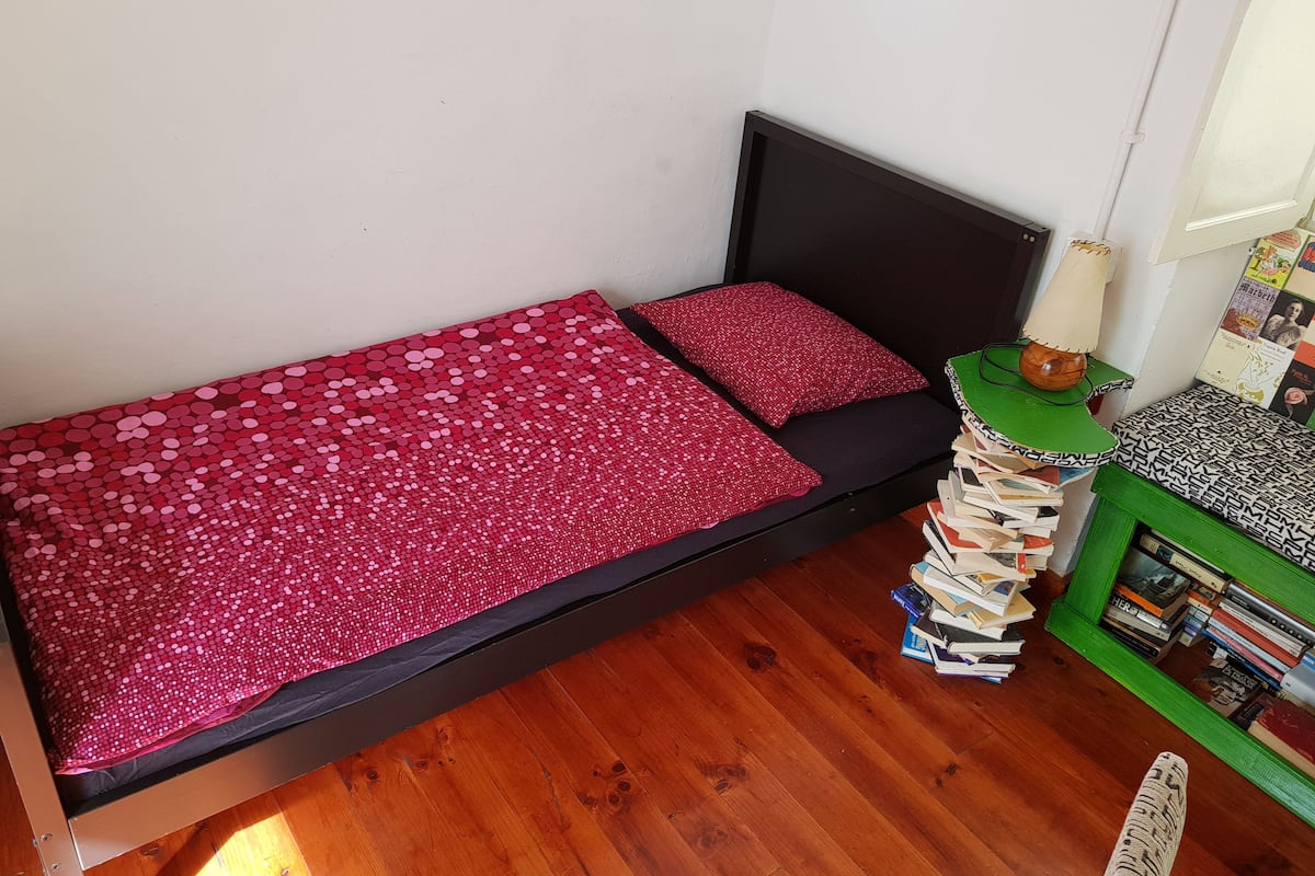 A small, single-bed room with a wooden floor, a black headboard, and a red sequin bedspread.