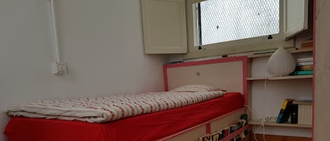 A single bed with a red mattress and white patterned bedding, a wooden headboard, and a shelf below filled with books and magazines.