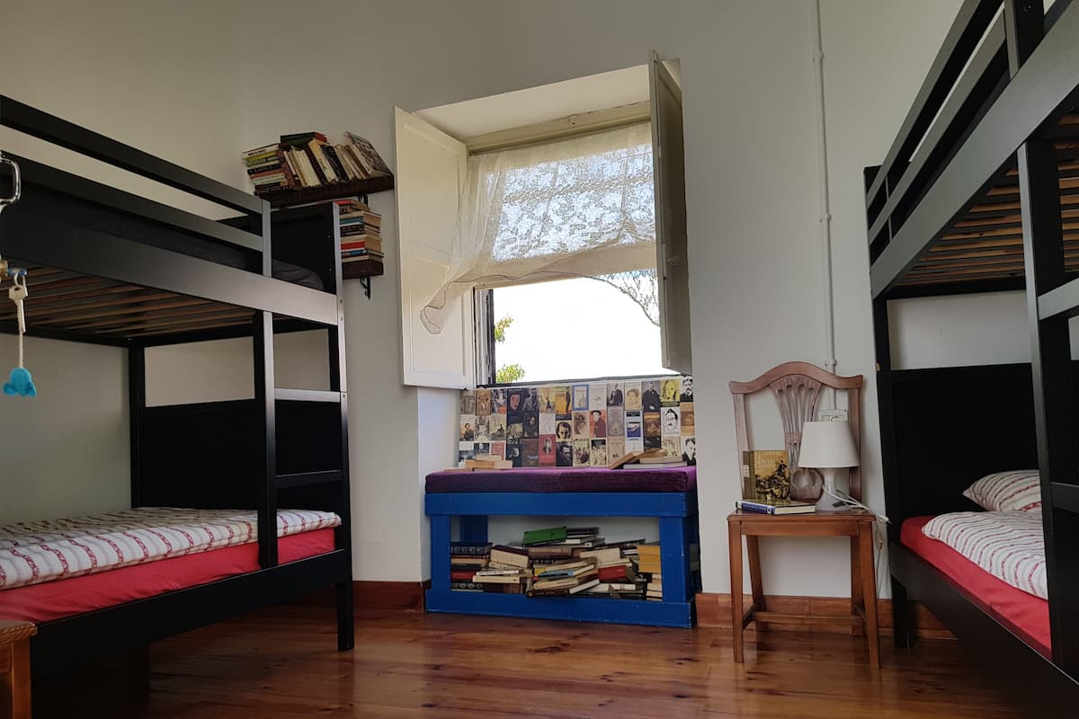 A dormitory room with bunk beds, a desk with books, a window, and a lamp.