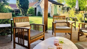 Une terrasse extérieure avec des chaises et une table en bois, entourée de verdure et avec un bâtiment en arrière-plan.