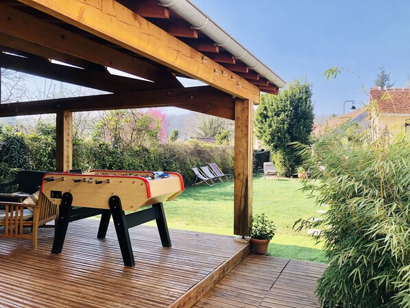 Una terraza de madera con mesa de fútbol de mesa, un área de estar cubierta y vistas a un jardín cuidado.
