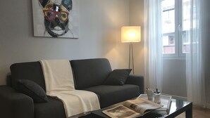 A modern living room with a dark grey sofa, a white throw blanket, and a glass coffee table with a magazine. A floor lamp and a window with curtains are also visible. - CENTER - SPACIOUS - COZY - WIFI (Albi)