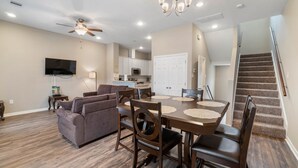 A modern living room with a sofa, dining table, and chairs. There is a ceiling fan, a flat-screen TV, and a kitchen in the background. - 10 mins from IAH / King Bed - Spacious 2 Bedroom home - Unit B (Humble)