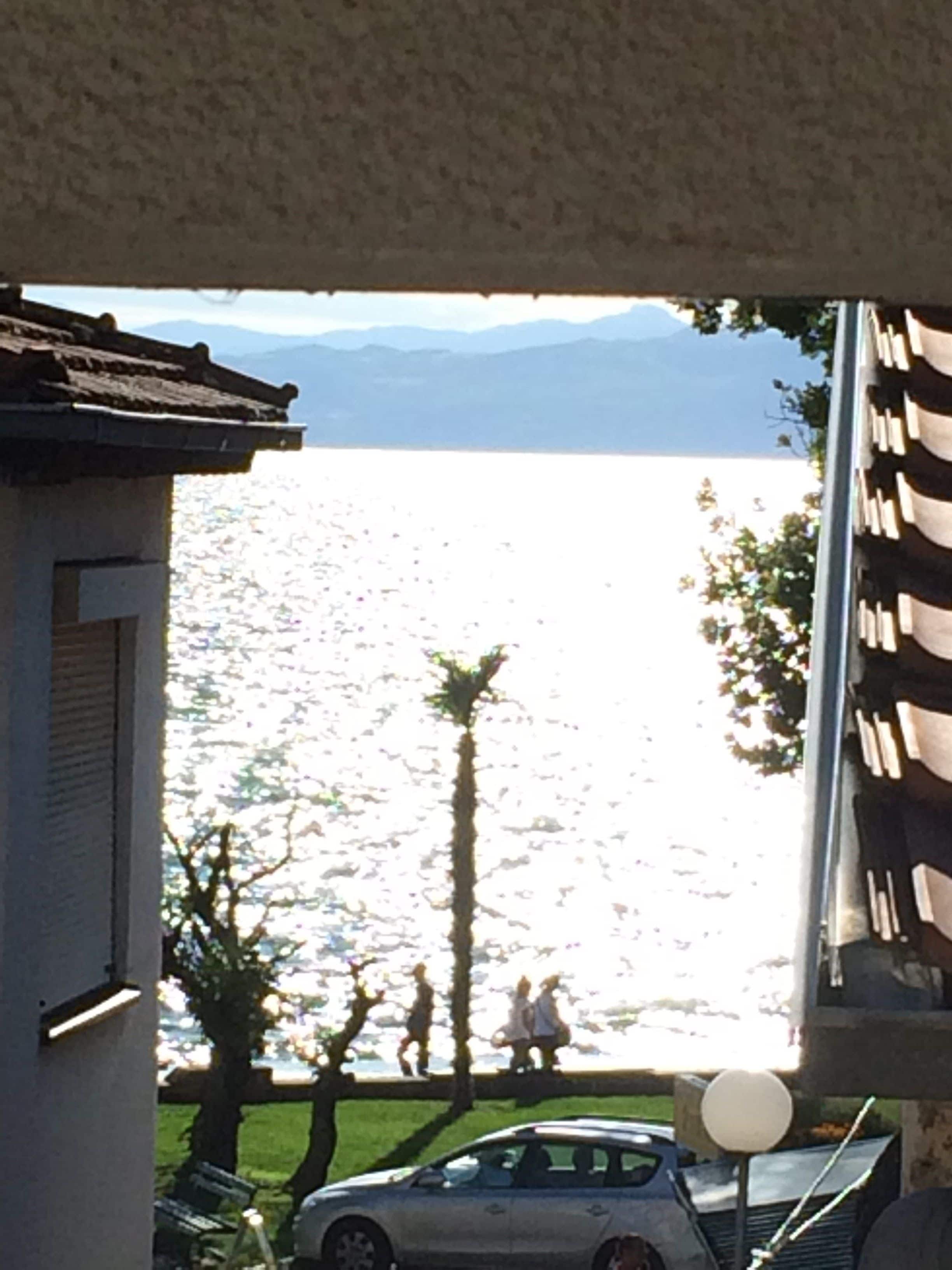 Vista a través de una ventana que muestra un lago y montañas al fondo. Hay un coche estacionado en primer plano.