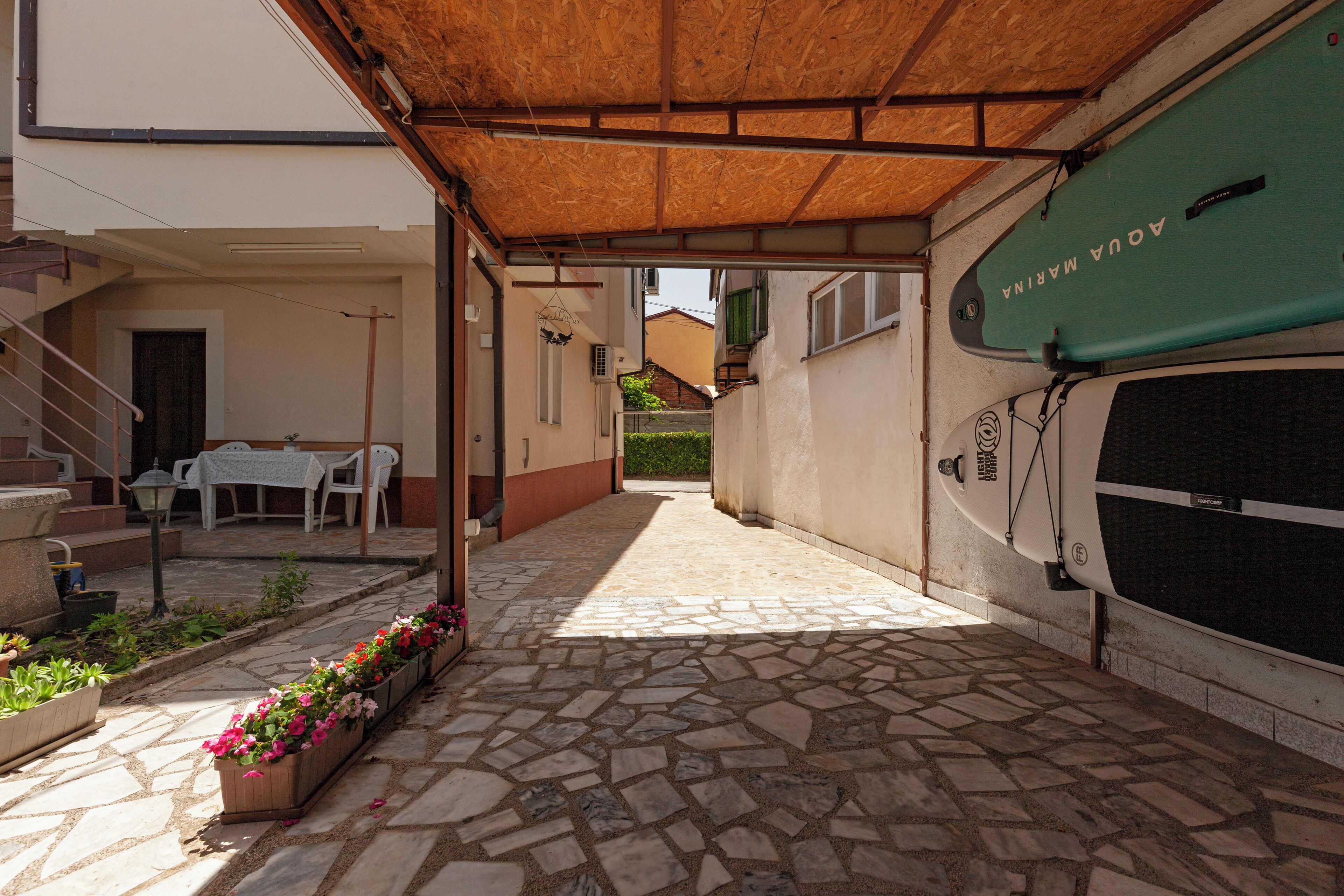 A covered outdoor area with stone flooring, a table with chairs, and a surfboard leaning against a wall.