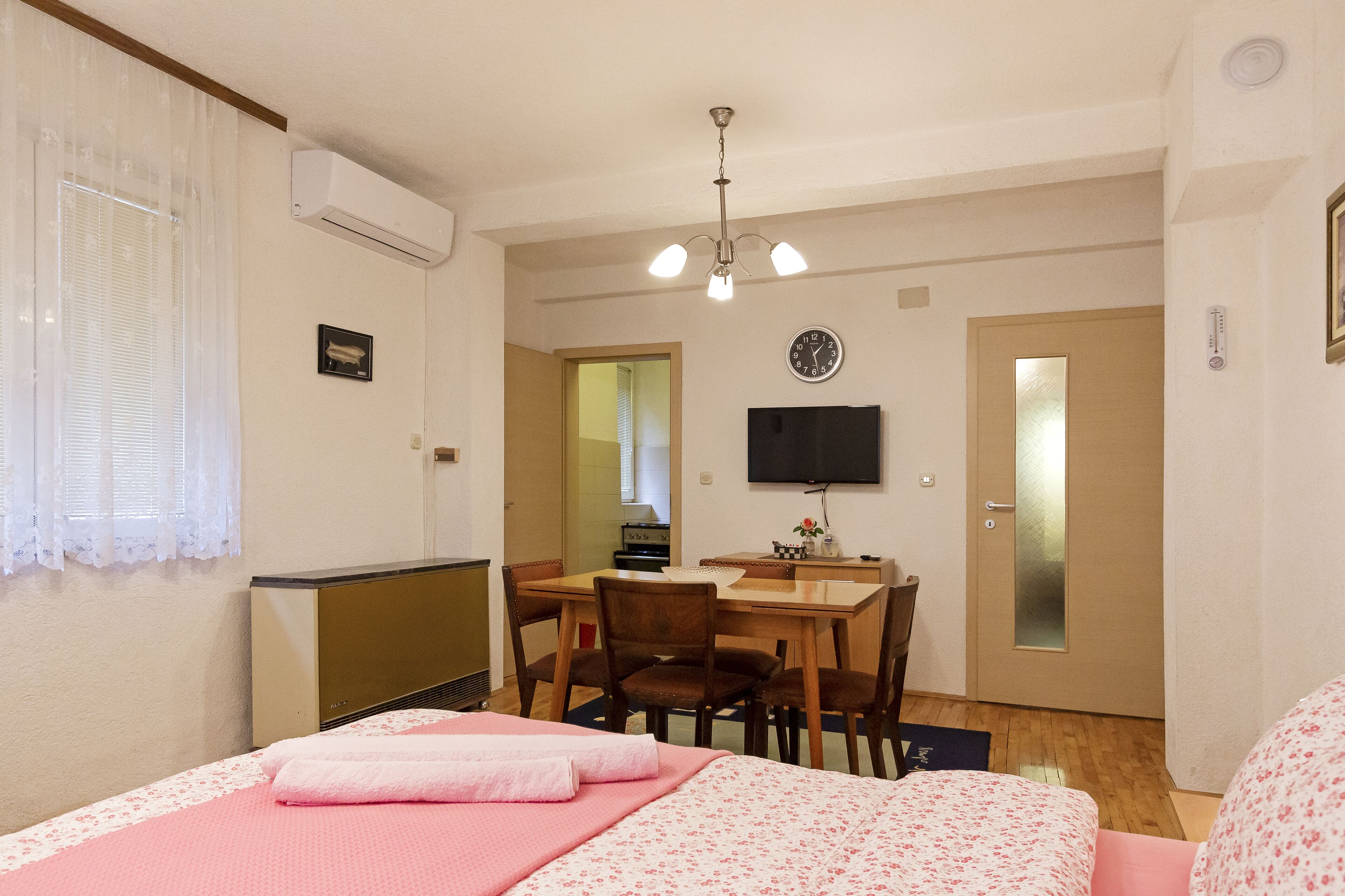 A bedroom with a bed, a desk, and a chair. There is a clock on the wall and a television mounted above the desk.