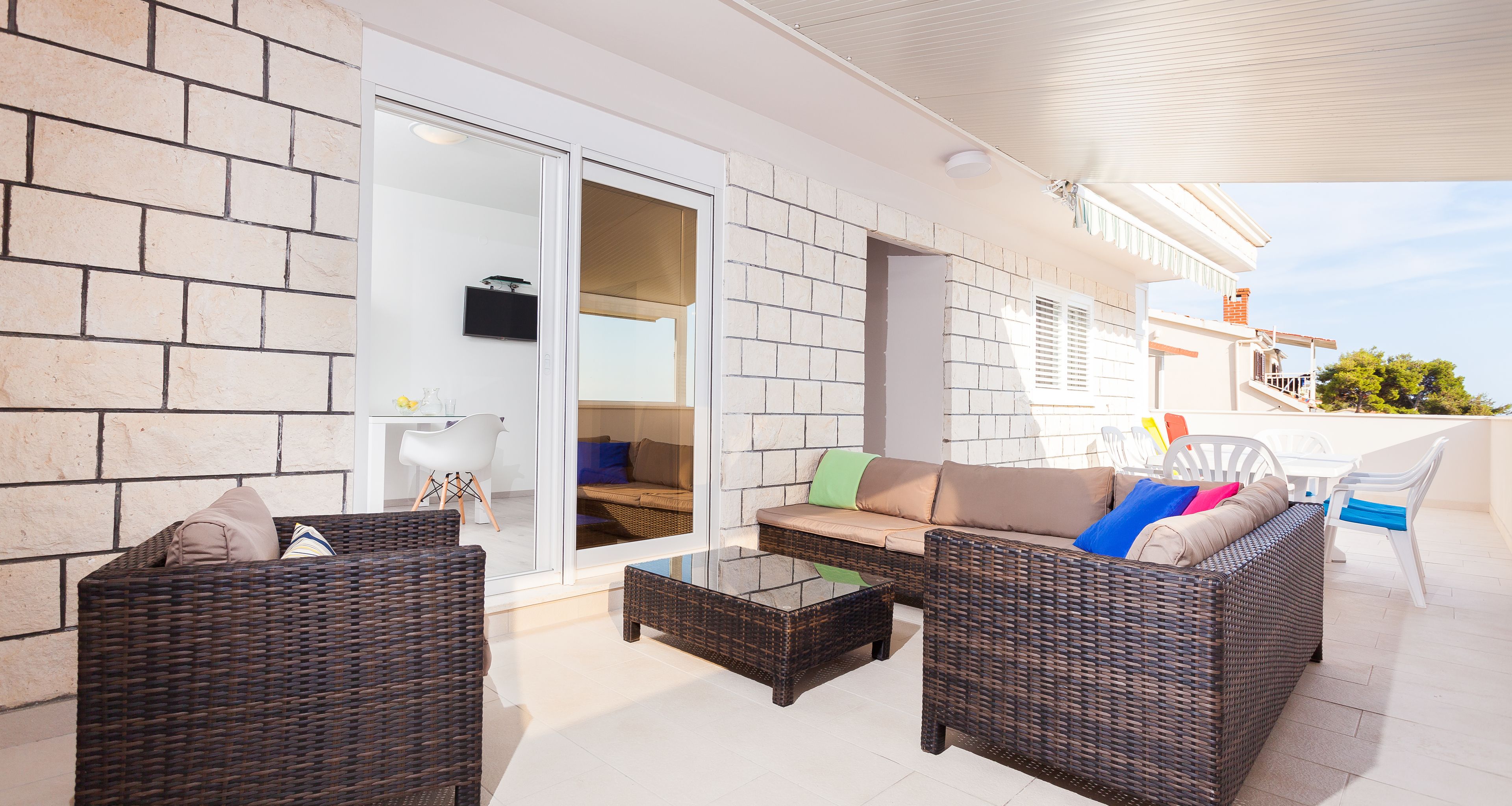 A covered outdoor seating area with wicker furniture, a glass table, and a white tiled floor.