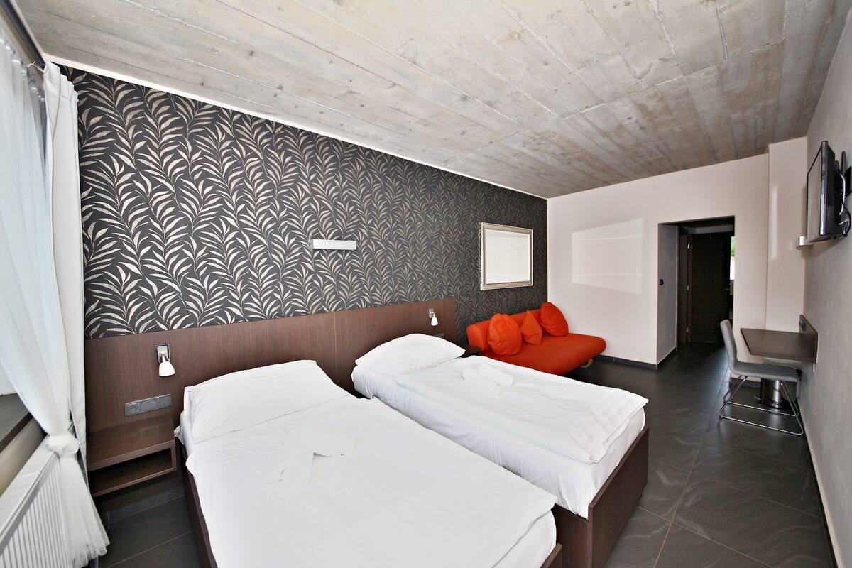 A modern hotel room with two beds, a wooden ceiling, a patterned wall, and a small seating area.
