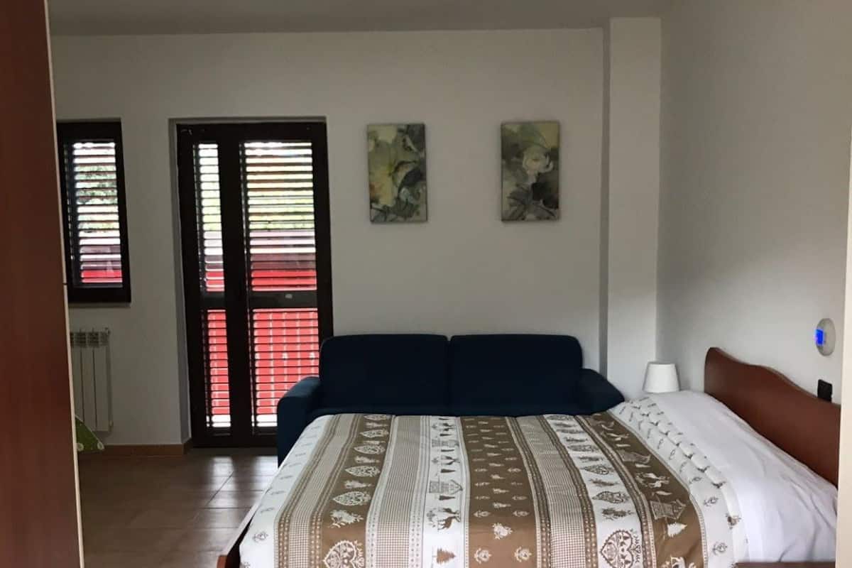 Quarto com cama, mesa de cabeceira, janela com persianas e porta.