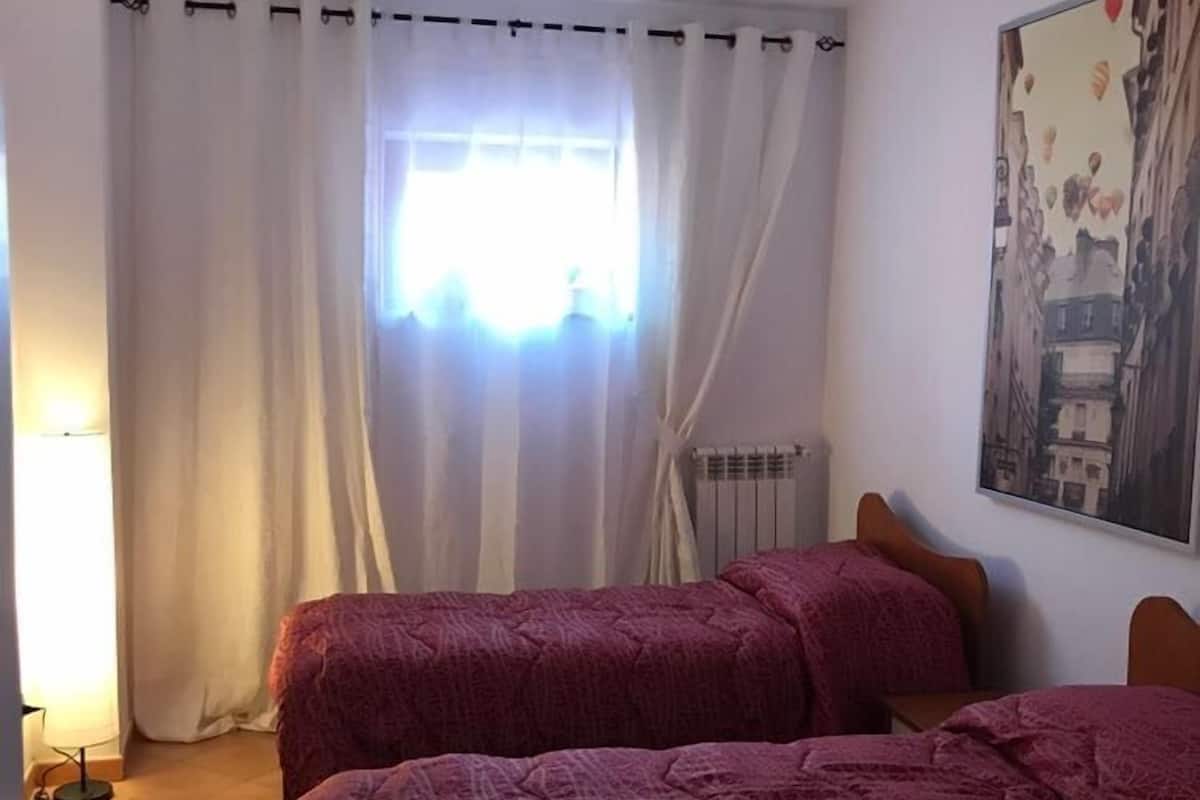 Quarto com duas camas, um radiador, janela com cortinas e um quadro na parede.