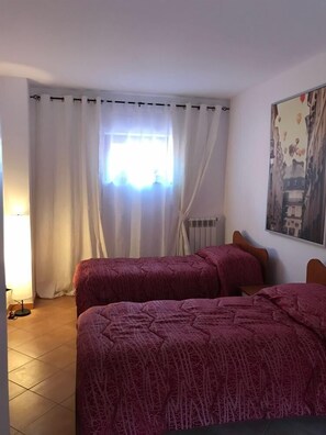 A room with two beds, a radiator, a window with curtains, and a painting on the wall.