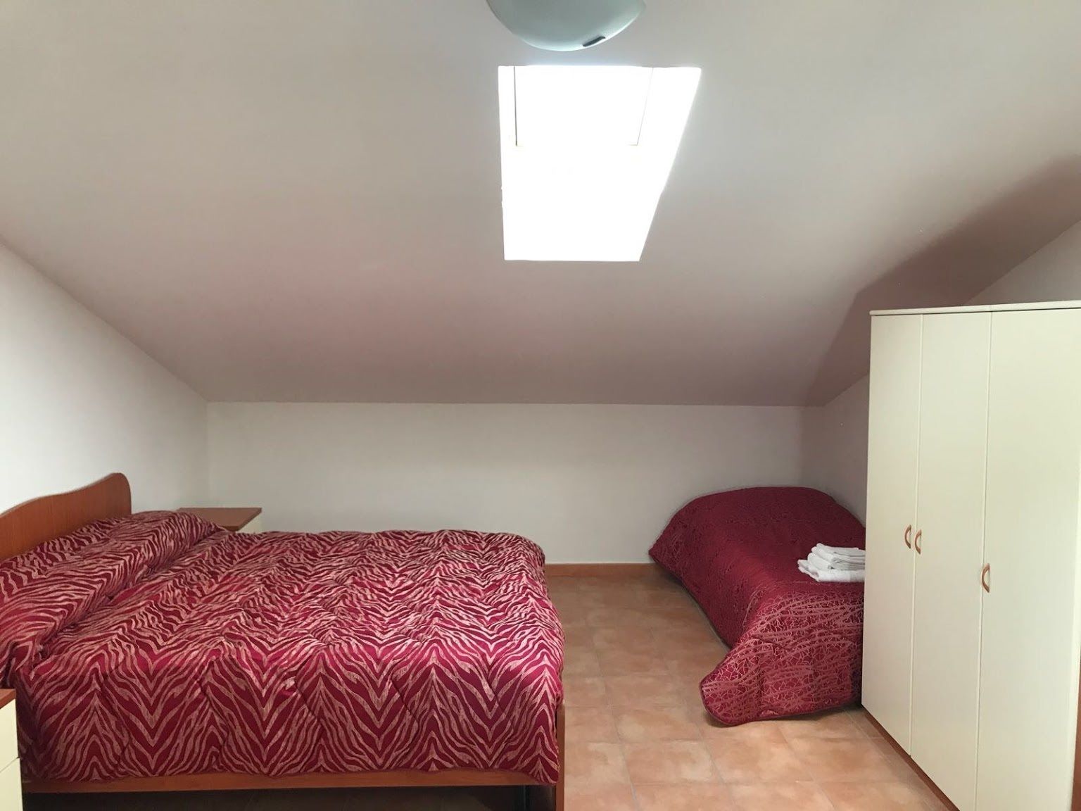 A small, simple room with a bed, a chair, a wardrobe, and a skylight.