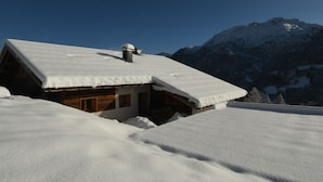 A snow-covered wooden cabin with a sloped roof, surrounded by snow-covered mountains and clear blue sky.