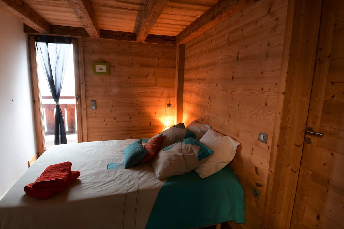A wooden cabin room with a bed, a window with curtains, and a wall-mounted light.
