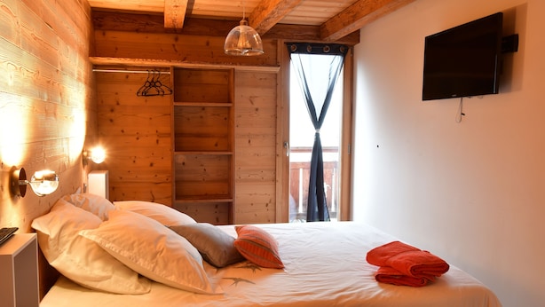 A wooden cabin room with a bed, a TV mounted on the wall, and a window with curtains.