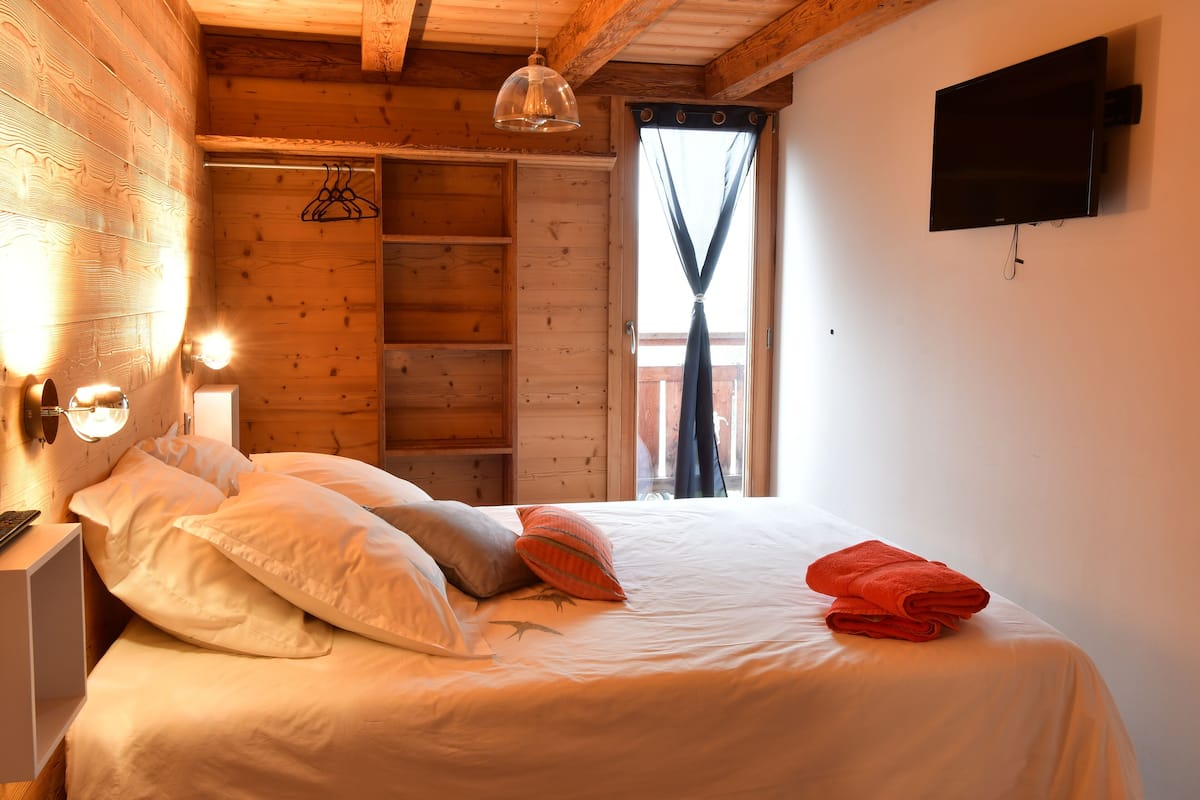 A wooden cabin room with a bed, a TV mounted on the wall, and a window with curtains.