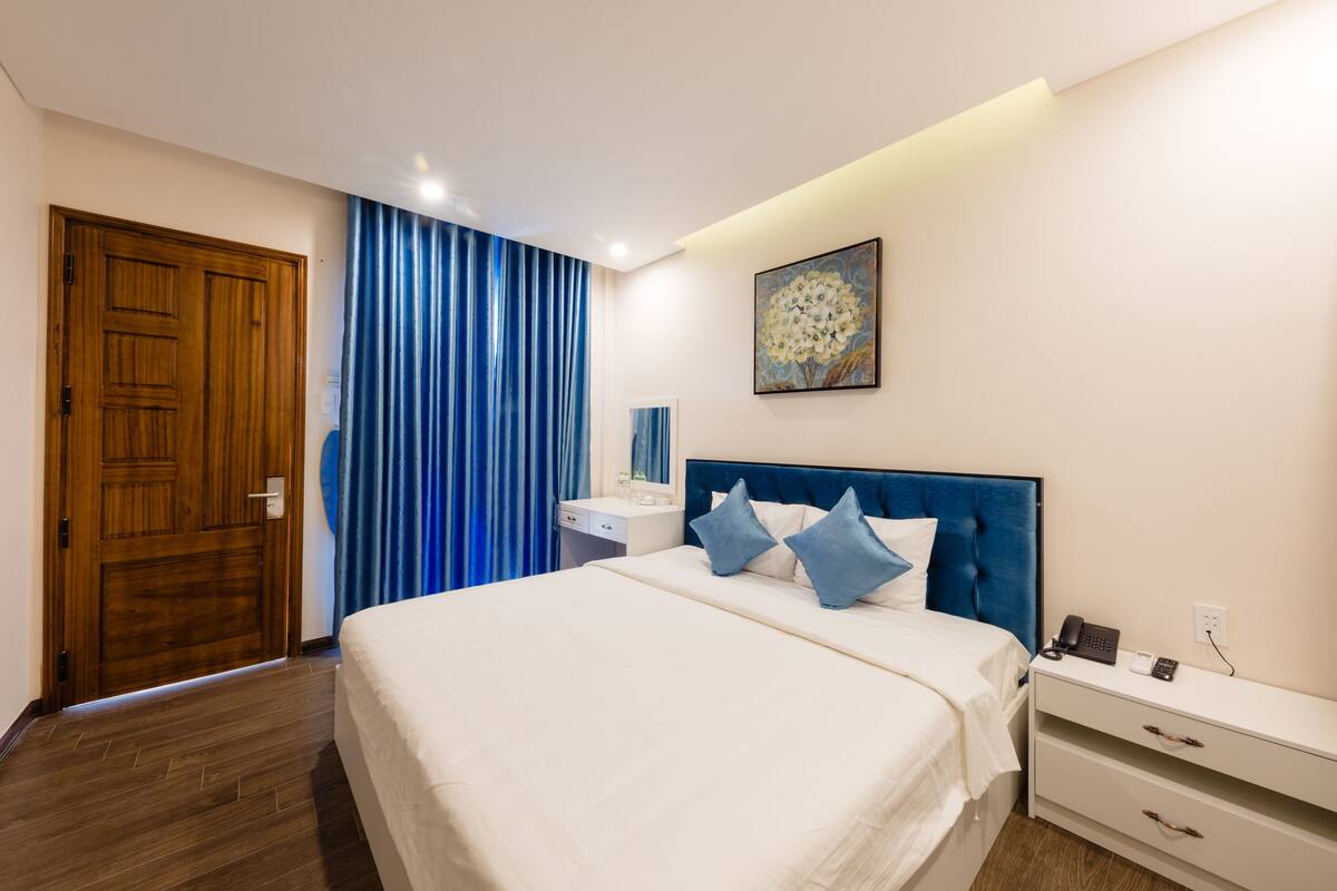 Una habitación de hotel con una cama grande, una mesita de noche con un teléfono, una mesita auxiliar y una puerta de madera, junto con cortinas azules.