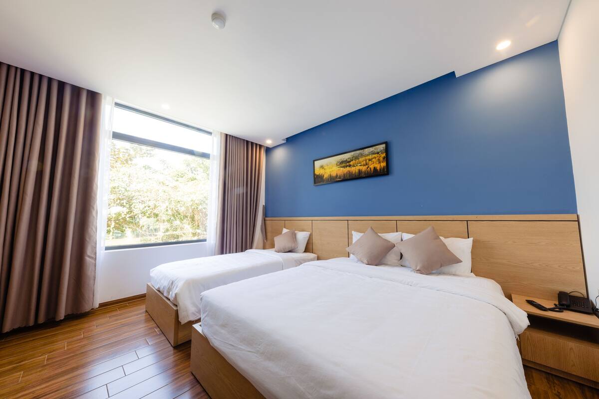 Una habitación de hotel con dos camas, suelo de madera, un ventanal con cortinas, una pared de acento azul y un cuadro.