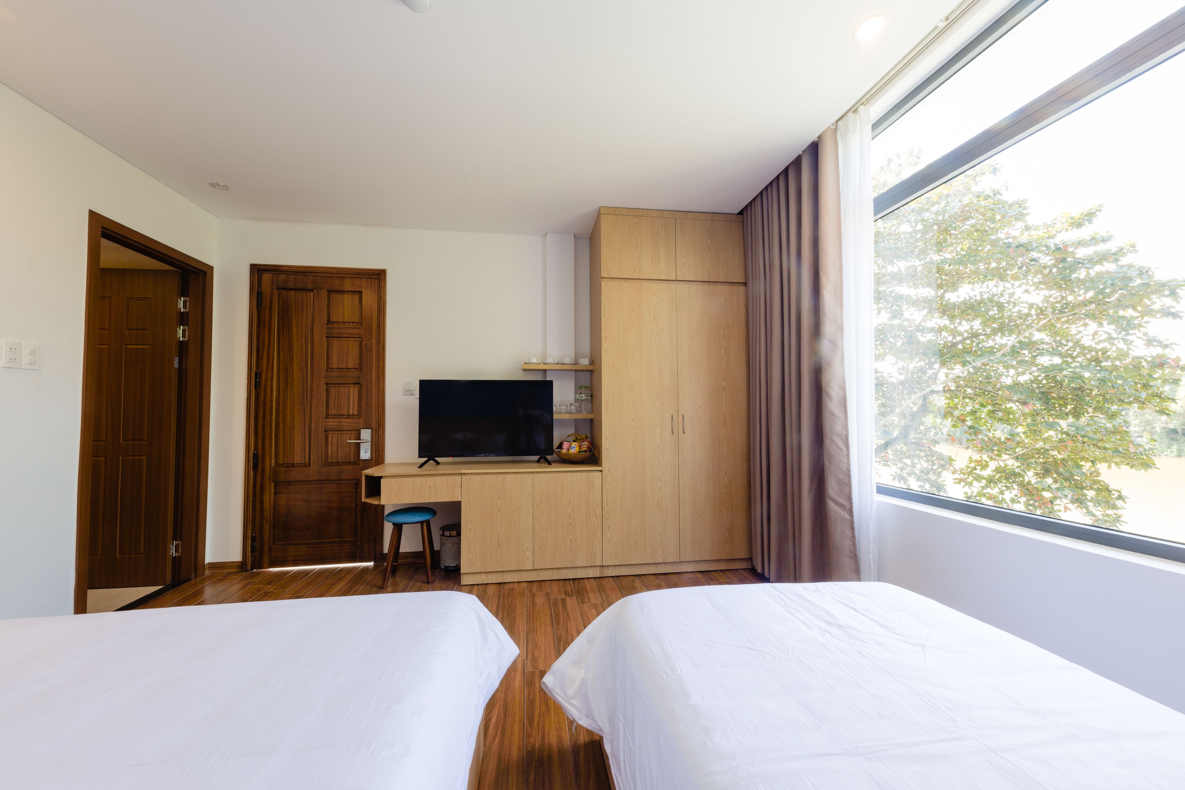 Quarto de hotel com duas camas, uma mesa de trabalho com uma TV, armários de madeira e uma janela com vista para árvores.