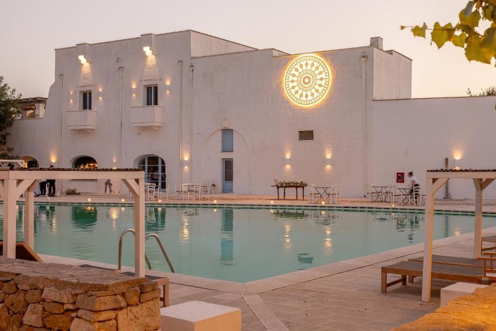 Zona de piscina con un edificio que tiene un diseño de luz circular en la fachada, mobiliario exterior y un muro de piedra.