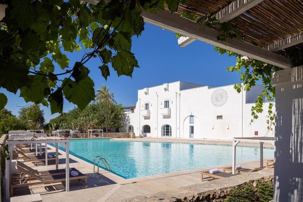 A swimming pool with lounge chairs, a white building with a circular window, and greenery in the foreground.